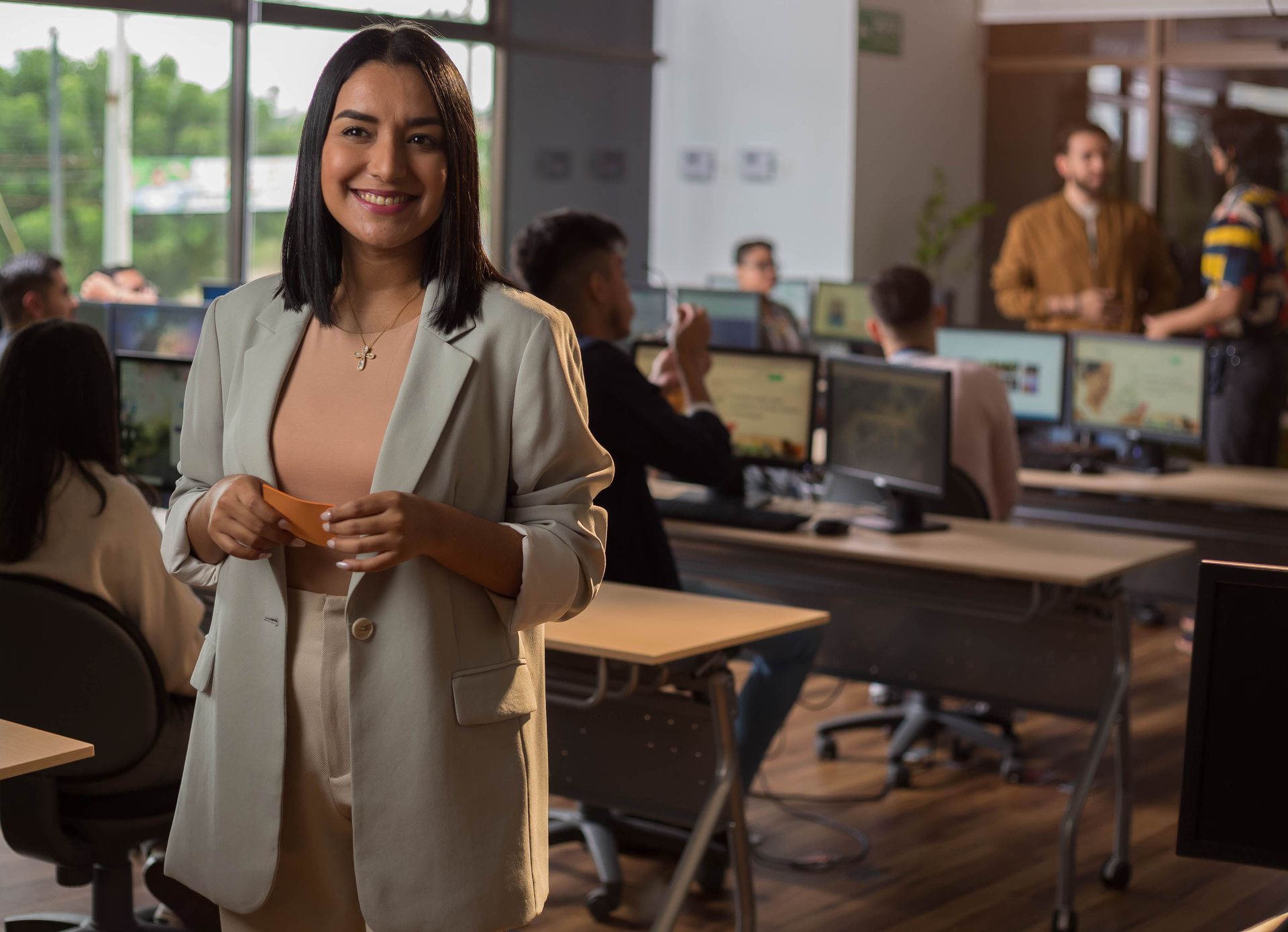 Smiling woman in a beige blazer standing in a modern office with coworkers at desks behind her