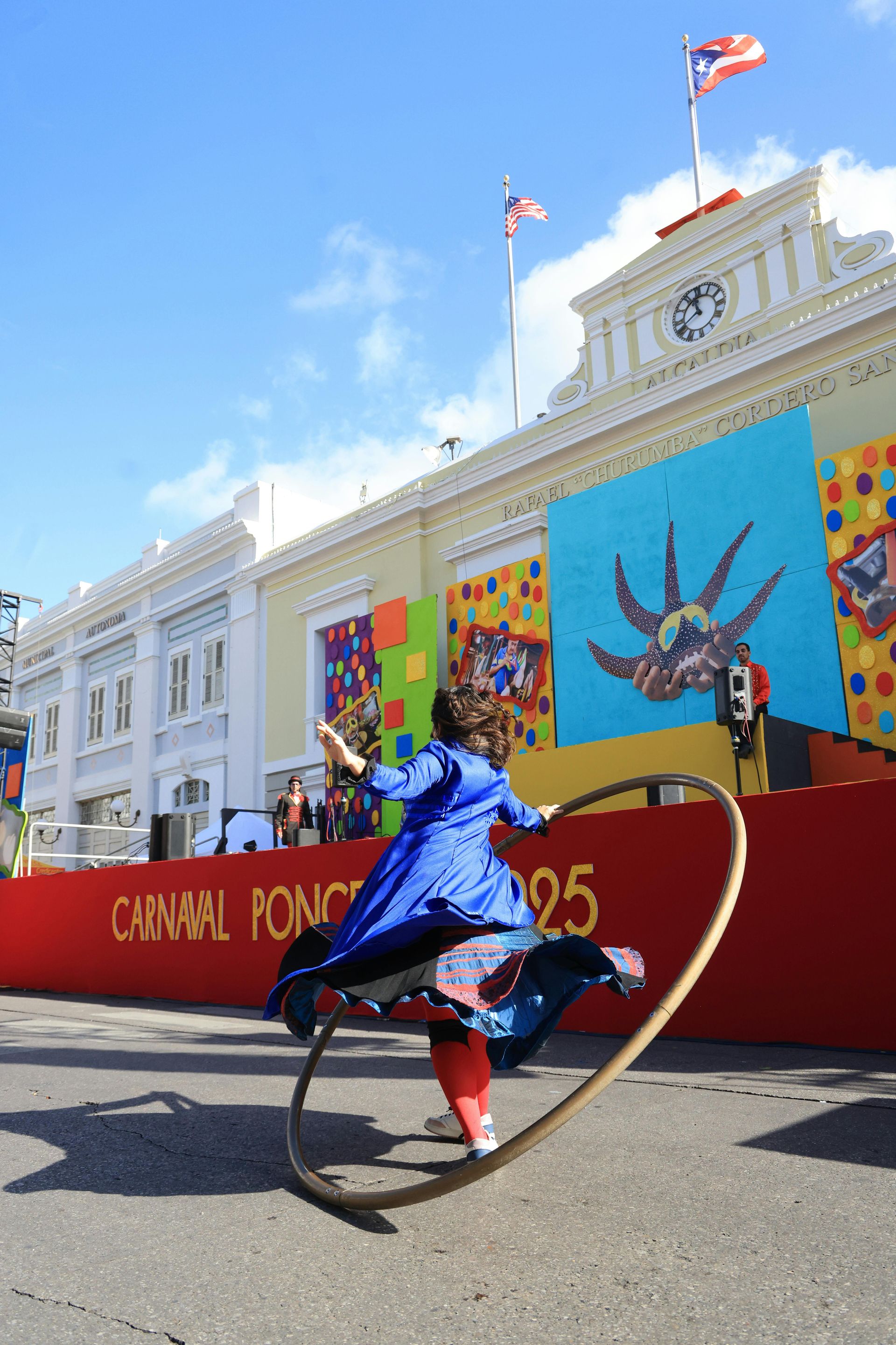 A performer in blue outfit with a hoop dances in front of a building with flags. Carnival banner is visible.