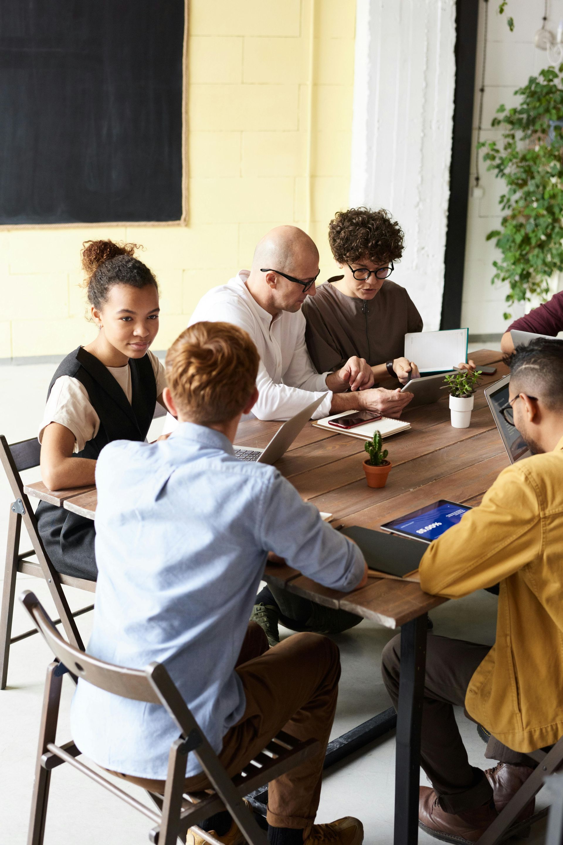 Five people seated around a wooden table in a bright office, collaborating on laptops and documents