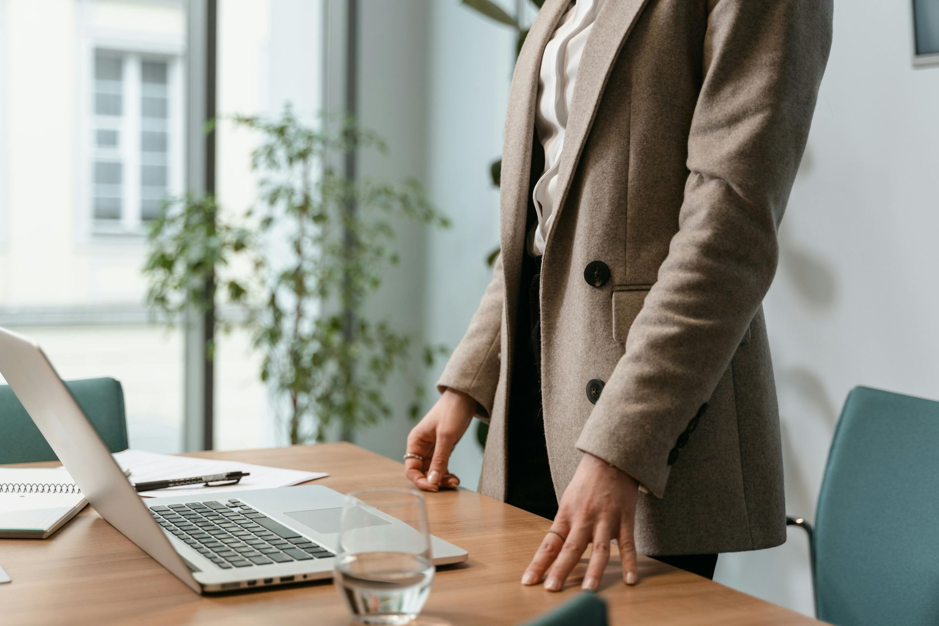 Woman in blazer stands by table with laptop and glass of water in office.