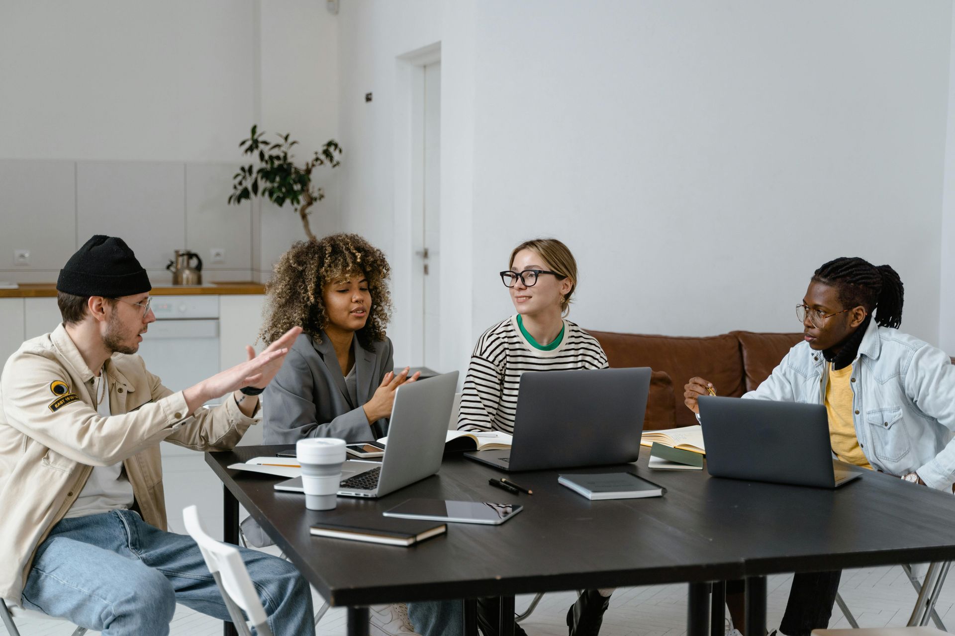 Four people in a meeting room discussing around a table with laptops and coffee cups.