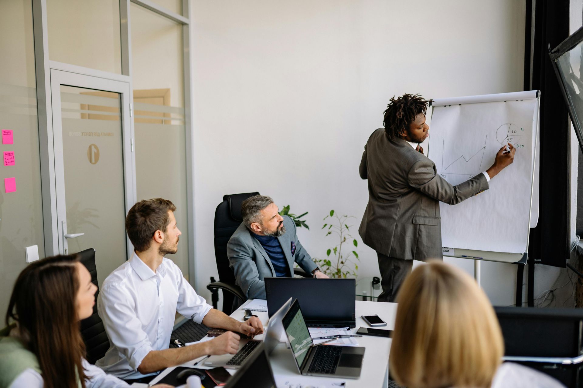 Team meeting in a bright office, presenter writing on a flip chart while colleagues watch around a table.