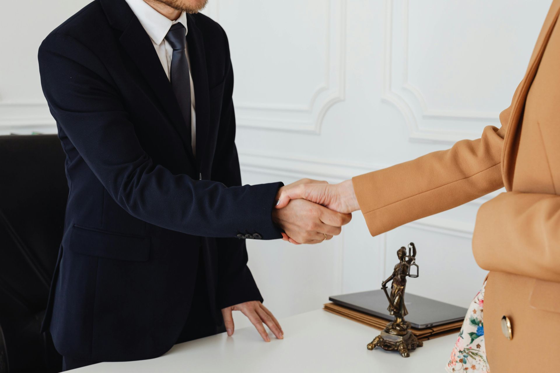 Two people in business attire shake hands over a desk; statue of Lady Justice in foreground.