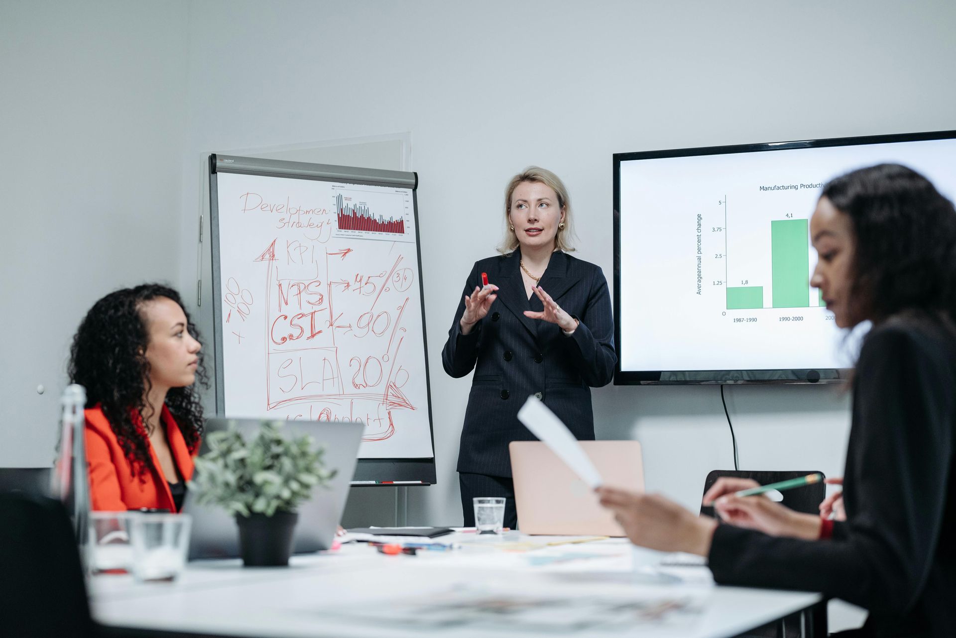 People in a conference room, listening to a presentation at a whiteboard. Natural light from a large window.