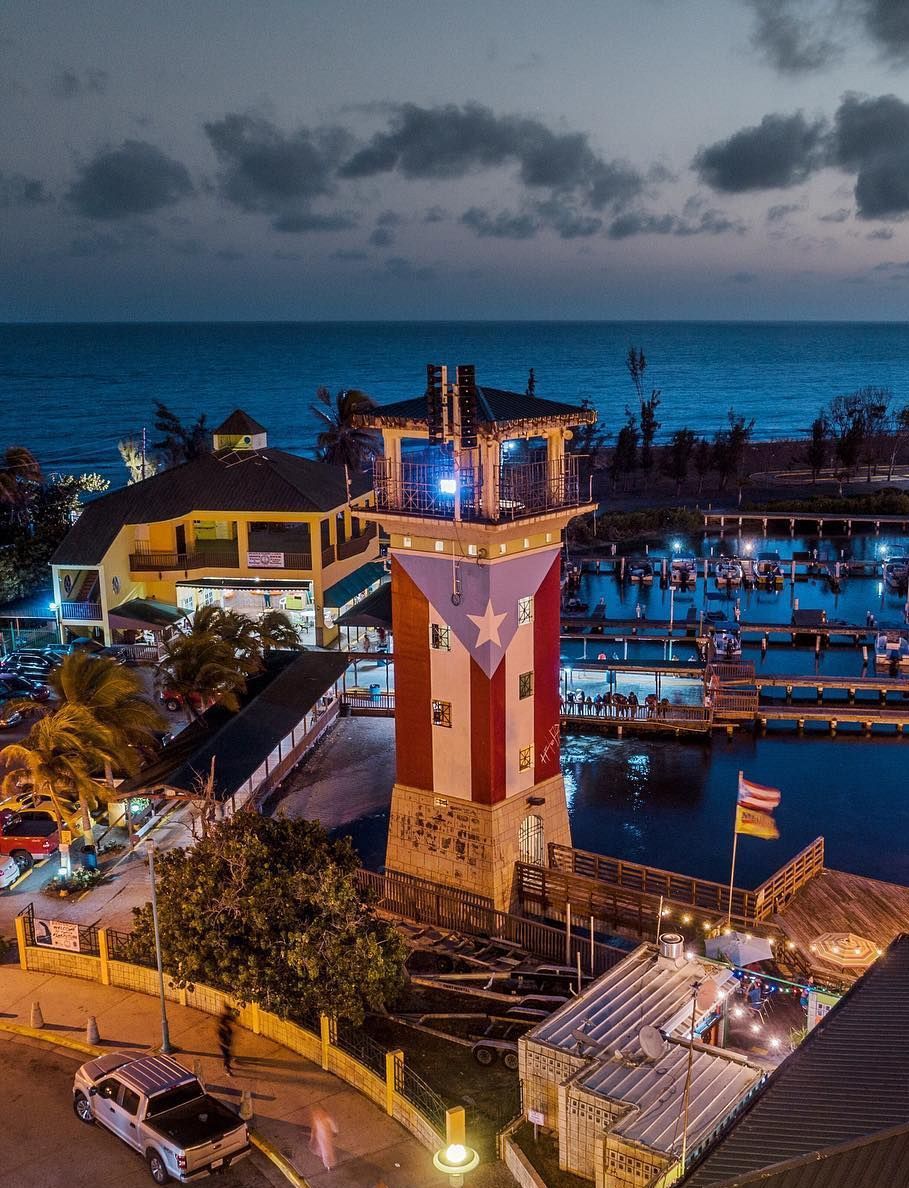 Lighthouse with the Puerto Rican flag painted on it, near a marina at dusk.