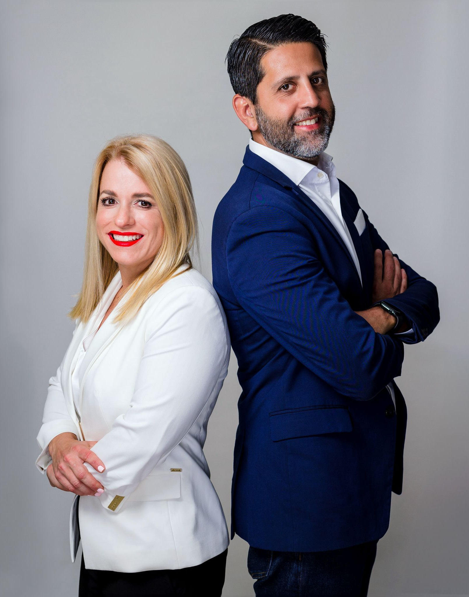 Marie and man Josean back-to-back, smiling. The woman wears a white blazer, the man, a blue jacket. Gray background.