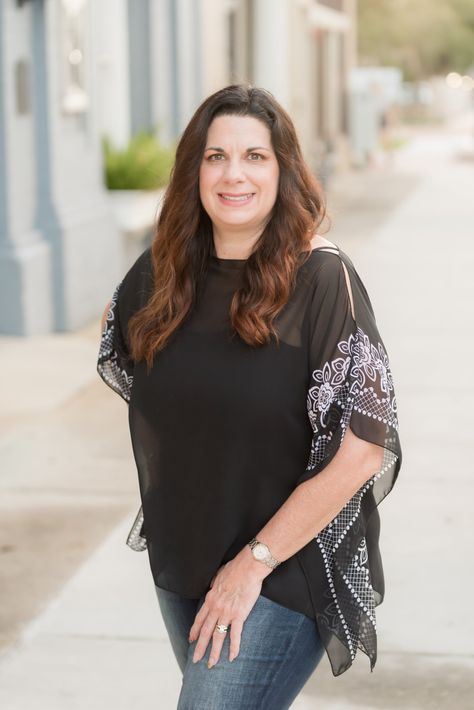 A person with long brown hair, wearing a black top with patterned sleeves and blue jeans, standing on a city sidewalk.