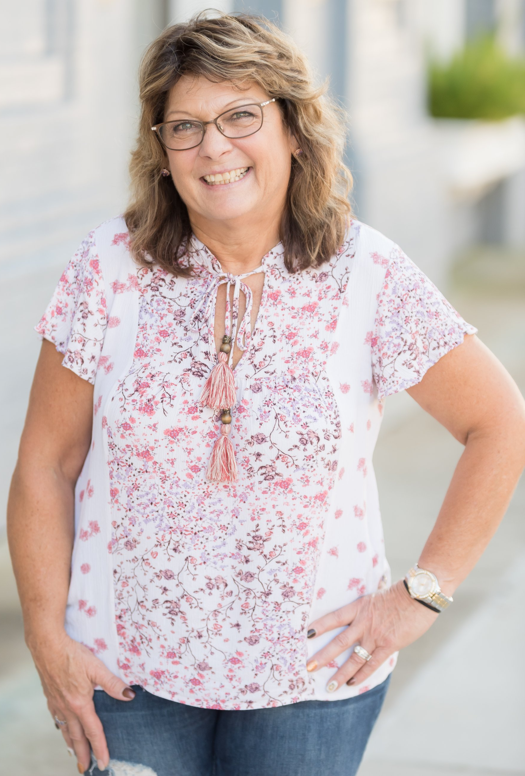 A smiling person wearing a floral white blouse and glasses, posing outdoors with a hand on their hip.
