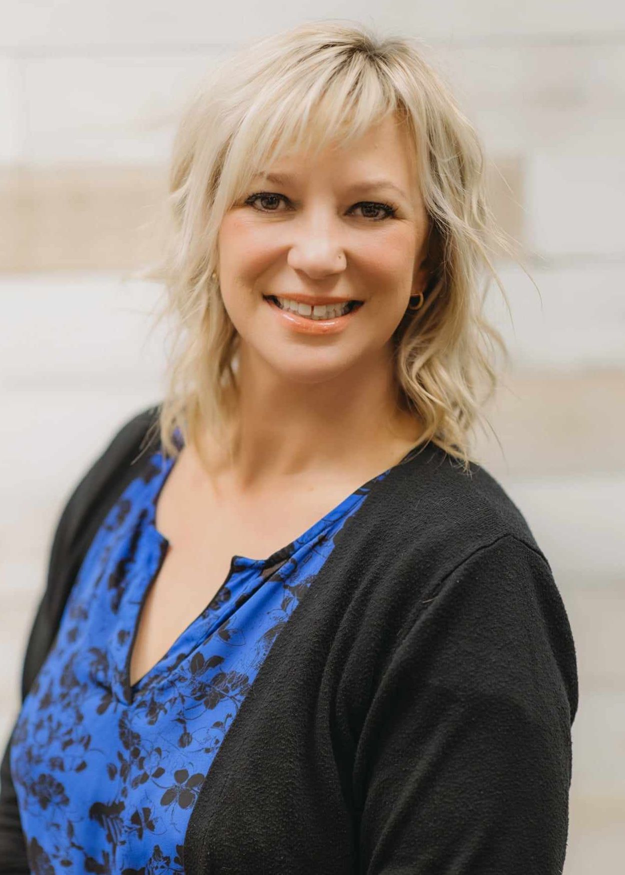 Headshot of a person with blonde hair smiling, wearing a blue floral patterned top and a black cardigan.