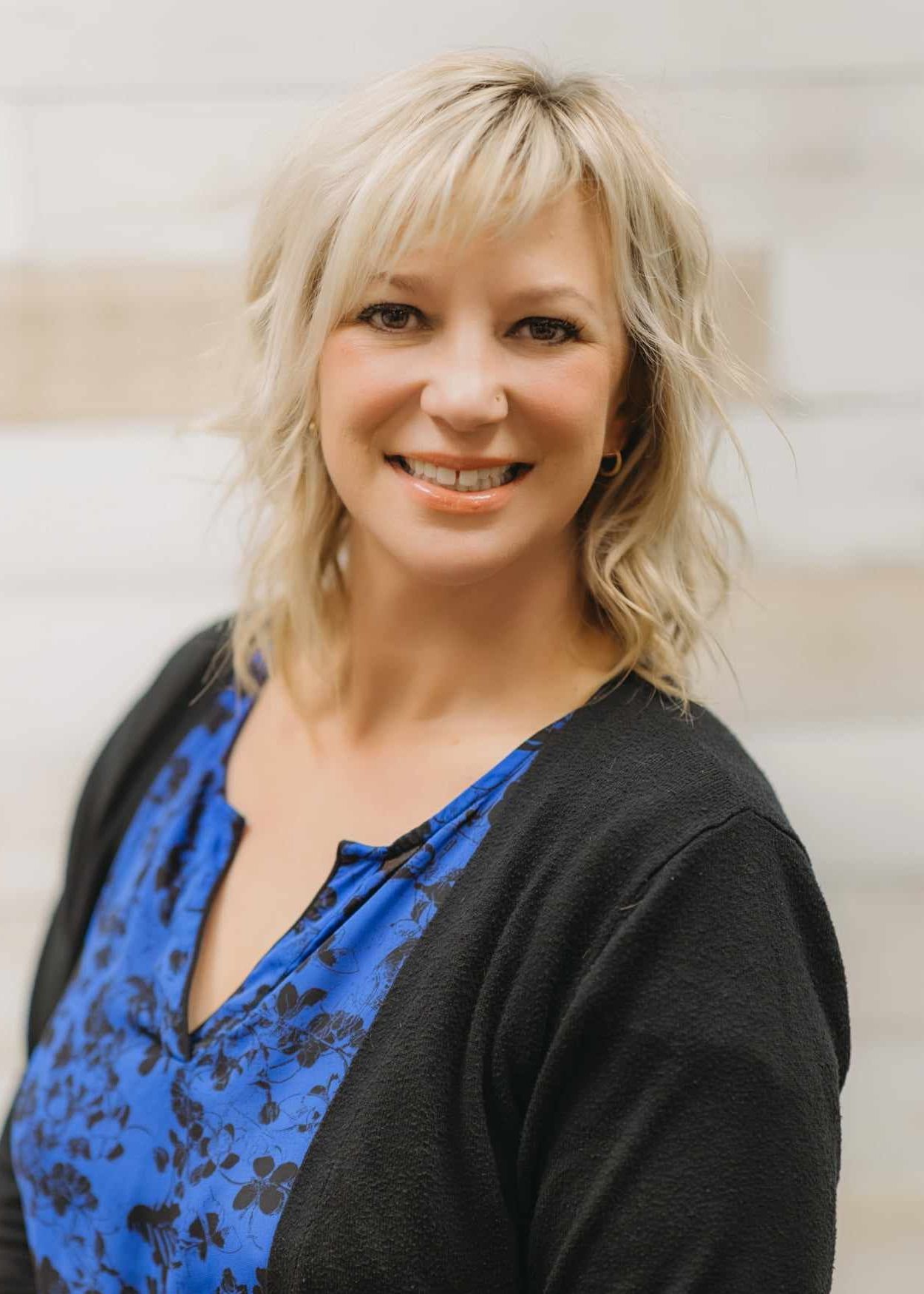 A smiling woman with blonde hair, wearing a blue floral top and a dark cardigan, against a light textured background.