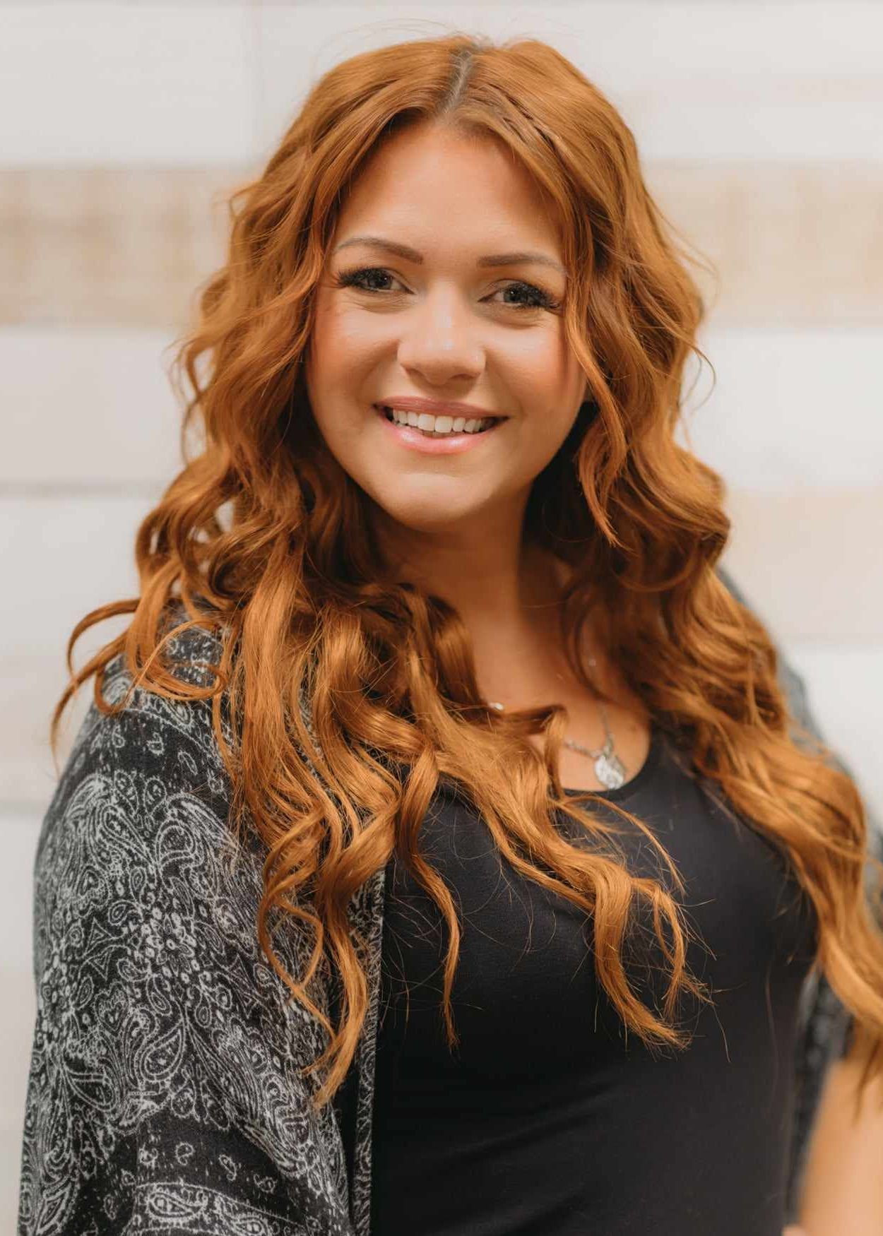 A smiling individual with long, wavy, auburn hair, wearing a black shirt and a patterned gray cardigan.
