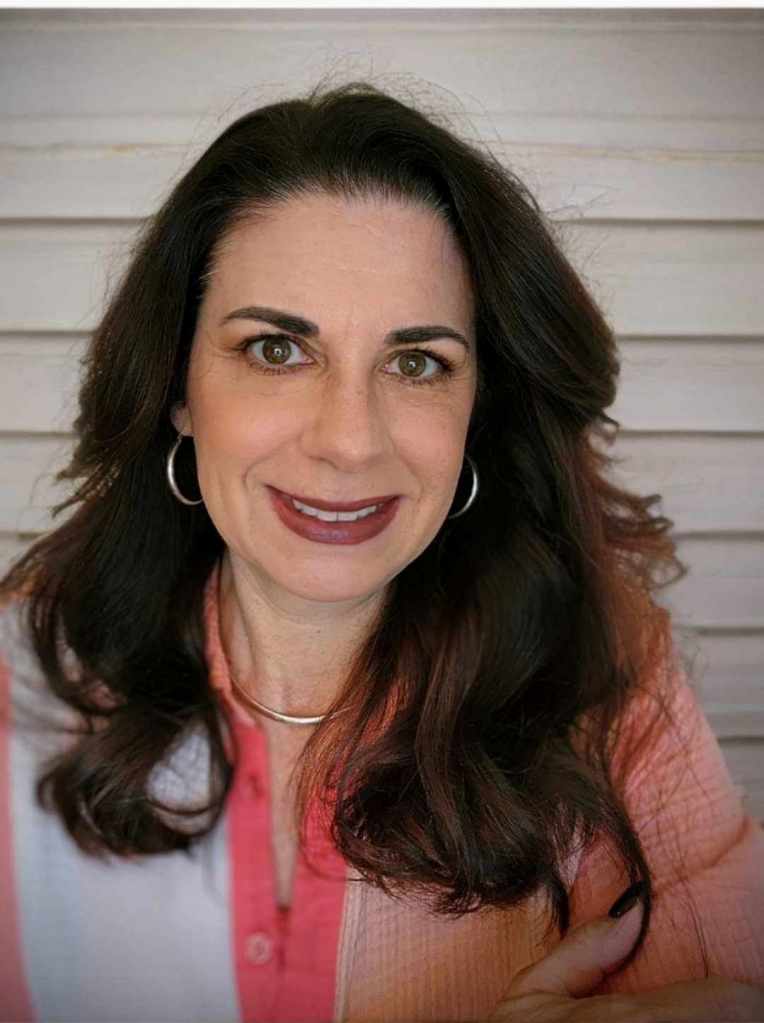 A person with long dark hair and silver hoop earrings wearing a peach button-down shirt, smiling against a white backdrop.