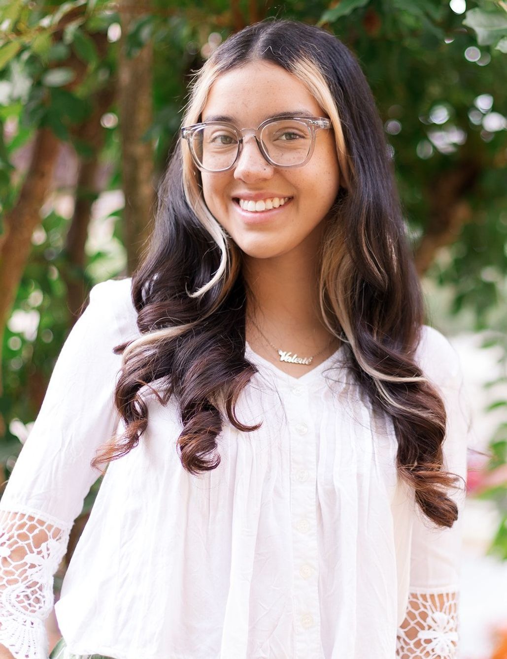 A smiling person with two-toned dark and blonde hair wears glasses and a white lace-trimmed top outdoors.