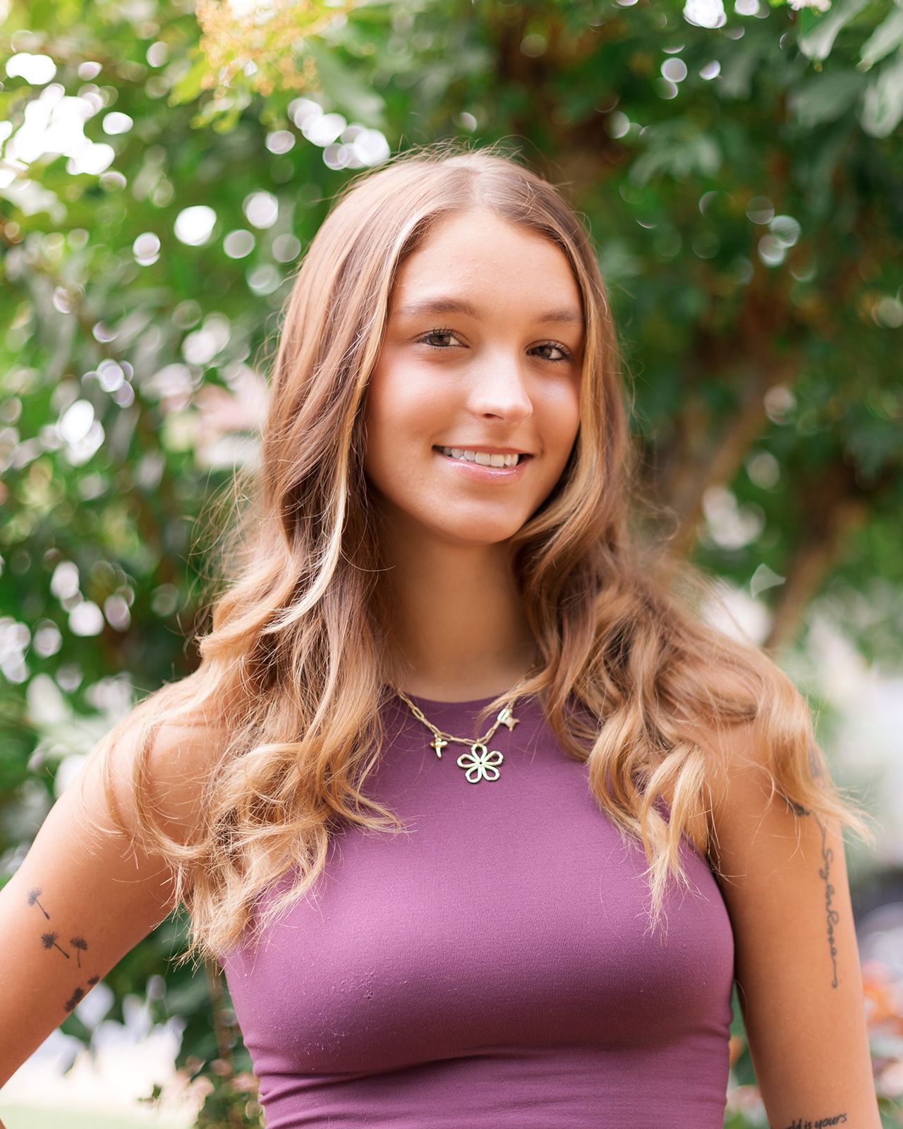 A smiling young person with long wavy hair wearing a mauve tank top and a flower-shaped necklace outdoors near trees.
