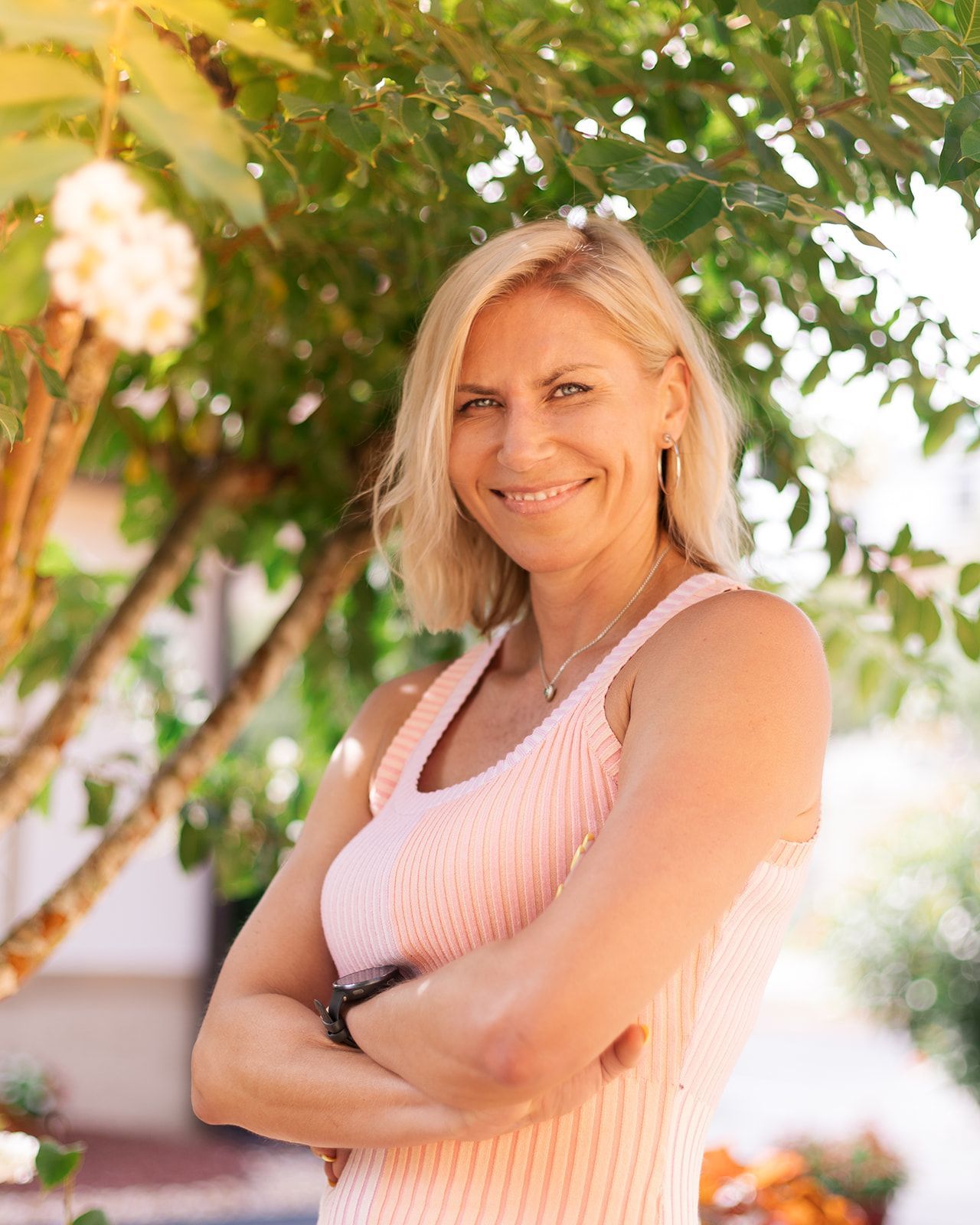 A smiling person with blonde hair stands with arms crossed outdoors, wearing a light pink top, set against leafy trees.