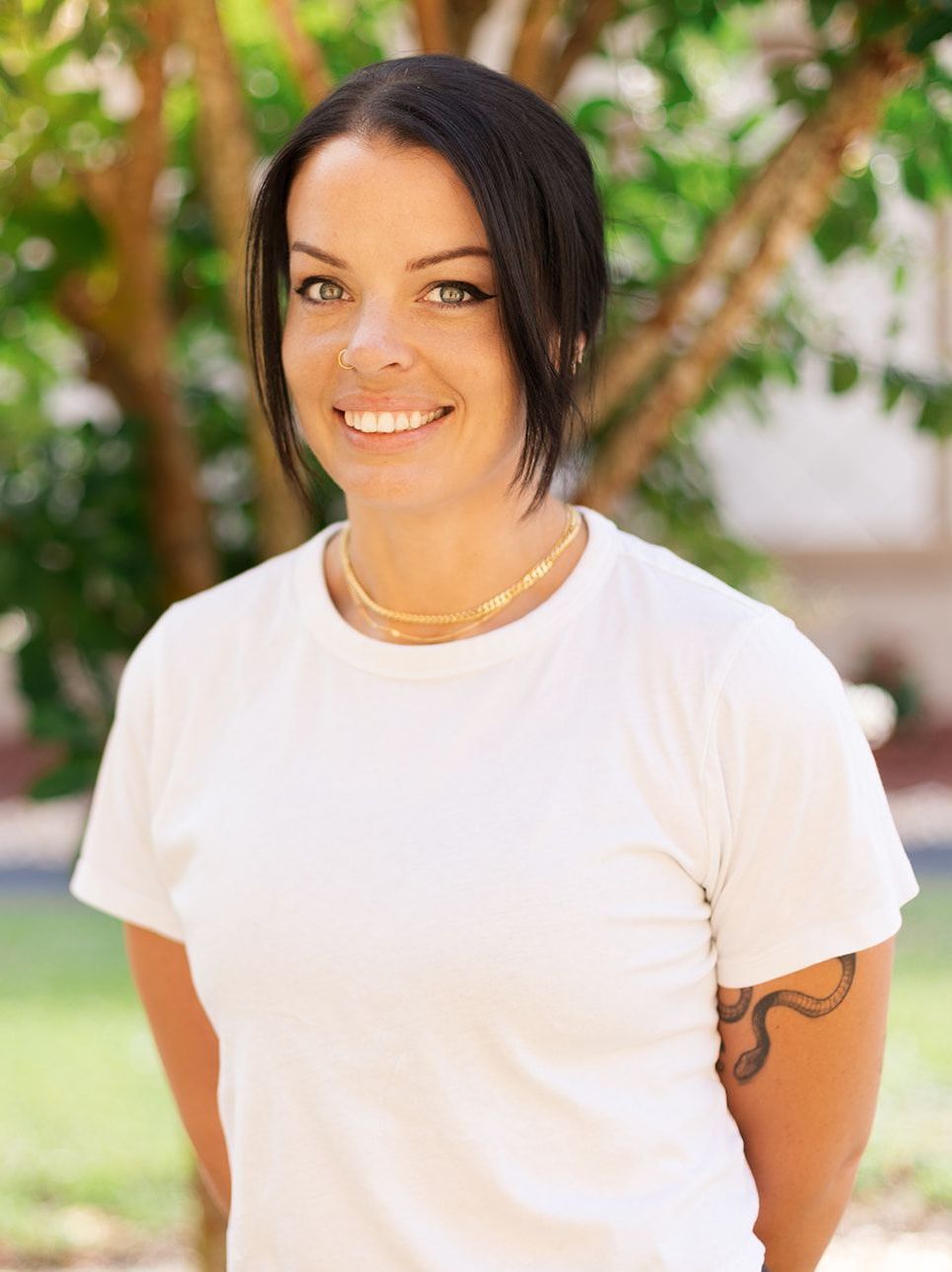 A person with dark hair, wearing a white t-shirt and gold chain, smiling in an outdoor setting with green foliage.