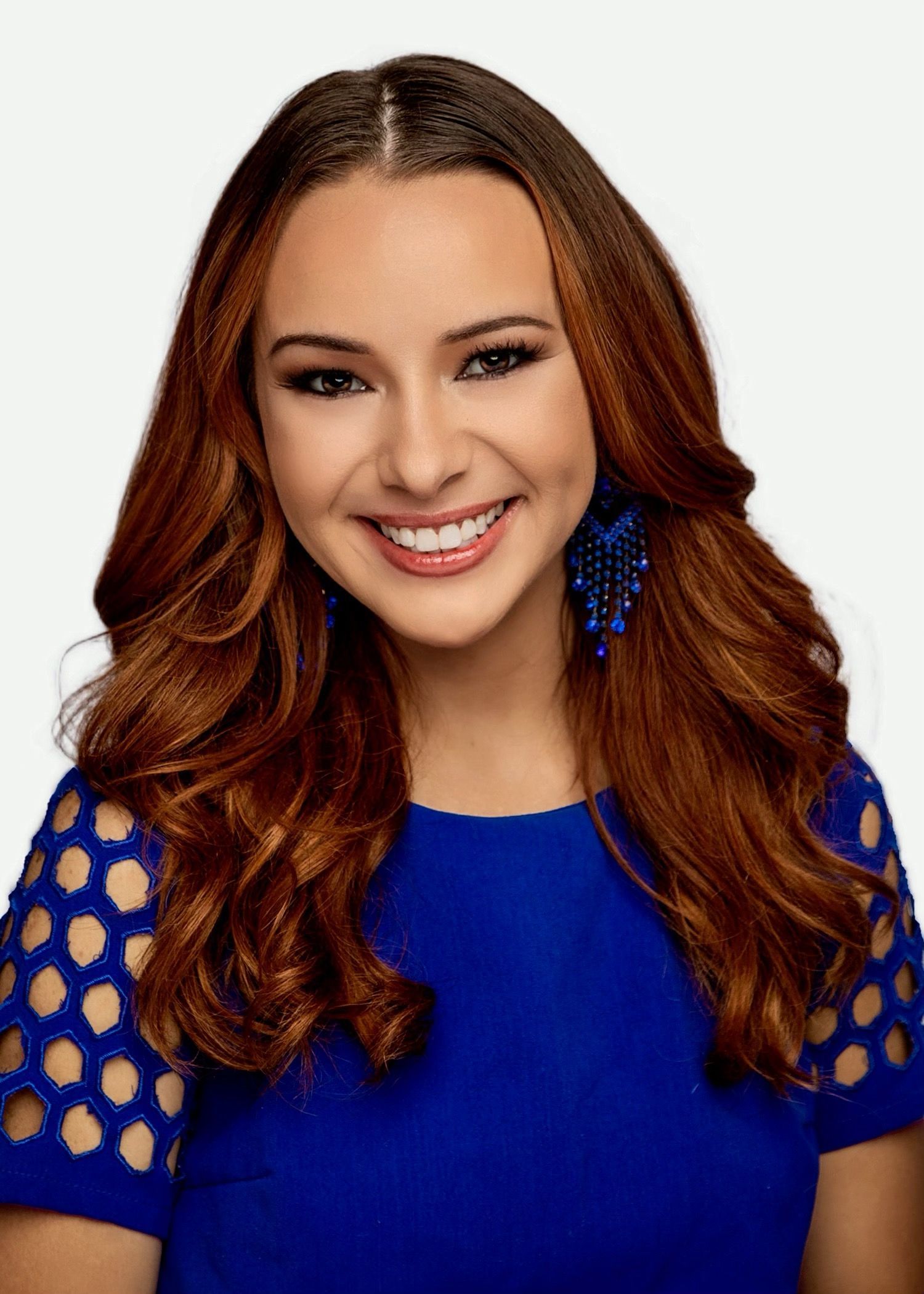 A smiling person with long brown hair, wearing a bright blue short-sleeved top with a circular cutout pattern and earrings.