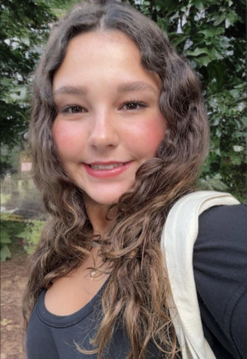 A person with long, wavy brown hair smiles at the camera, wearing a black top with a white bag strap, outdoors.