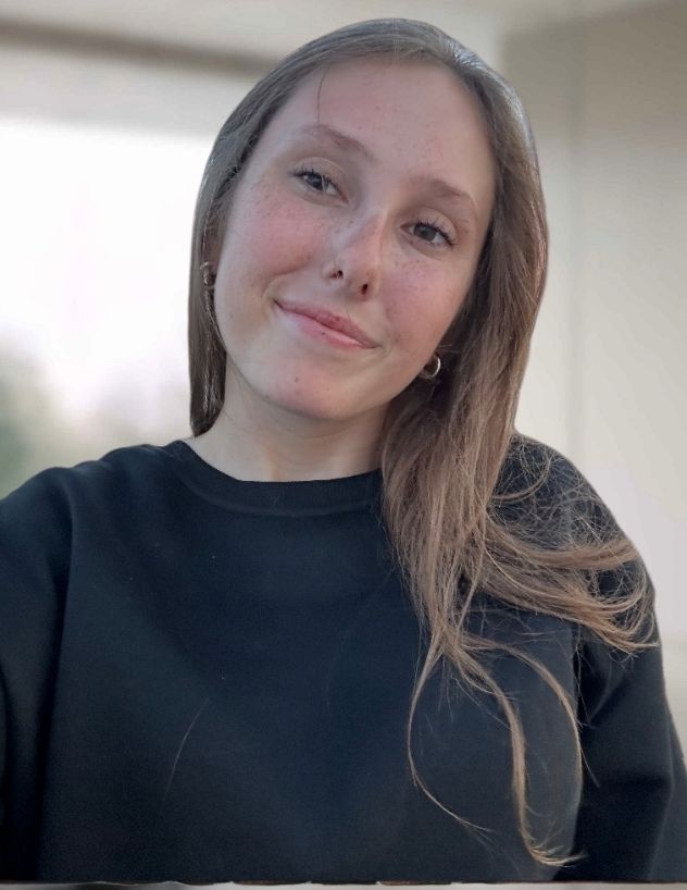 A person with long brown hair in a black sweater looking at the camera with a slight, gentle smile.