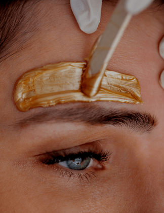 A close-up of a wooden spatula applying warm, gold-colored wax to a person's eyebrow area for hair removal.
