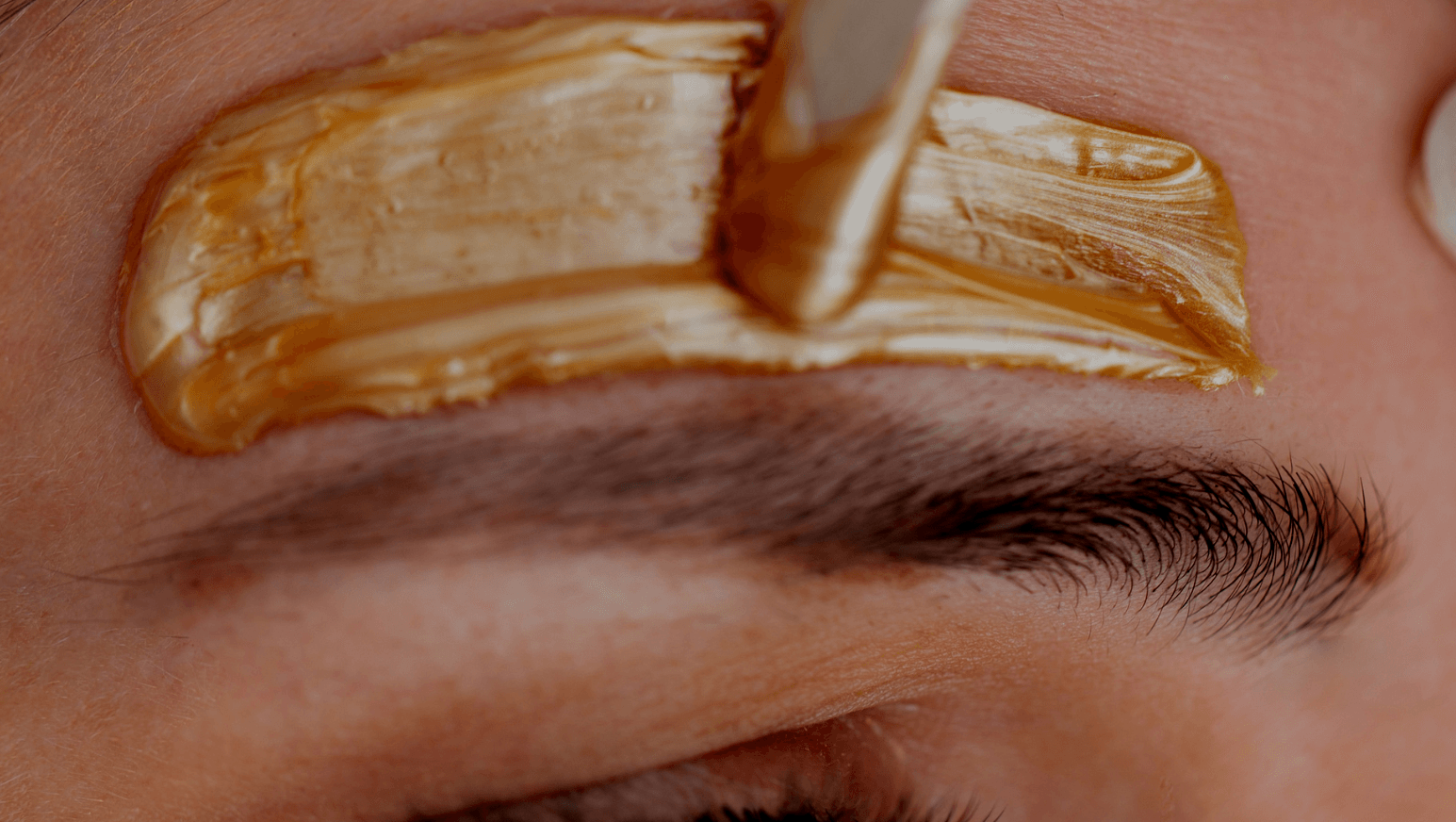A wooden applicator spreads warm, golden-colored hair removal wax onto the skin above an eyebrow.