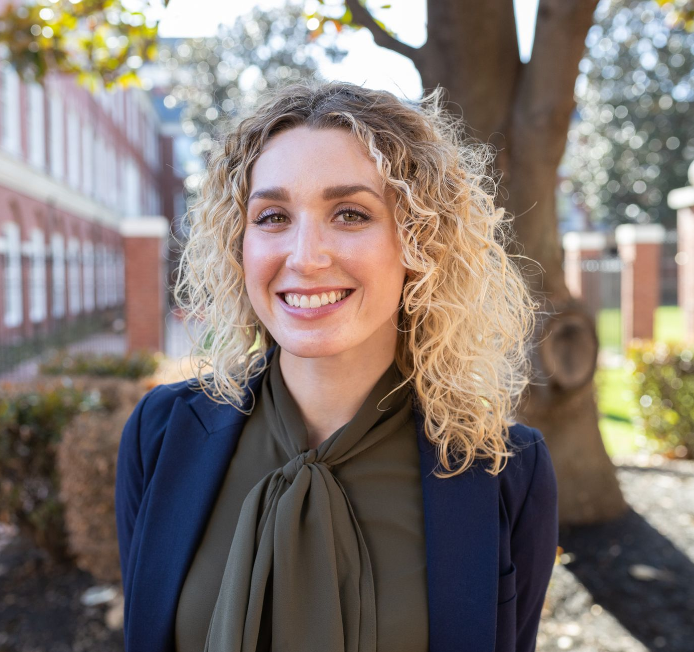 Woman in a navy blazer and green blouse smiles in front of a brick building and a tree.