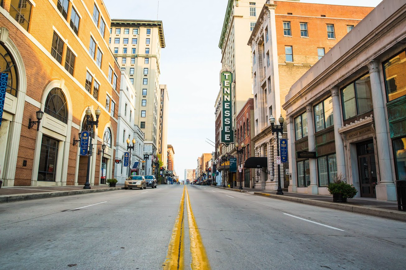 Empty street in a city, flanked by multi-story brick and stone buildings under a bright sky.