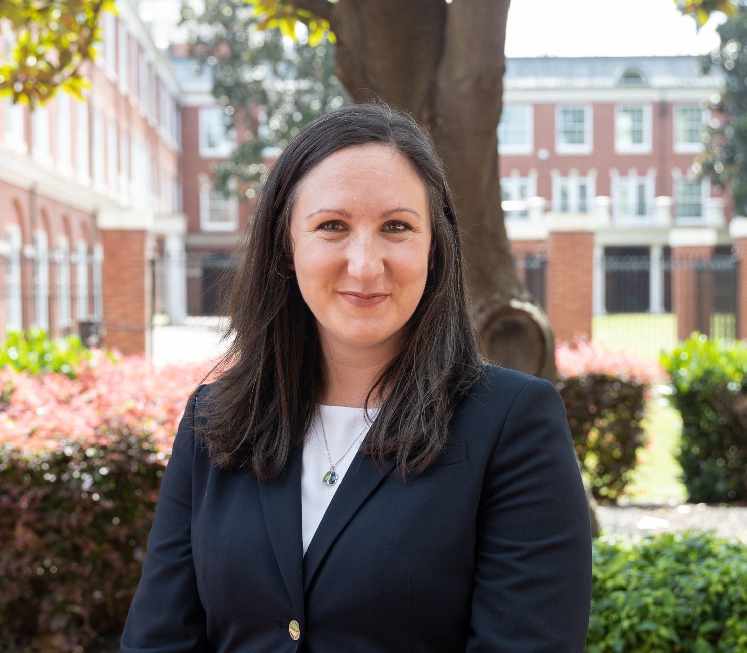 Woman in a blazer smiles in front of a brick building and foliage.