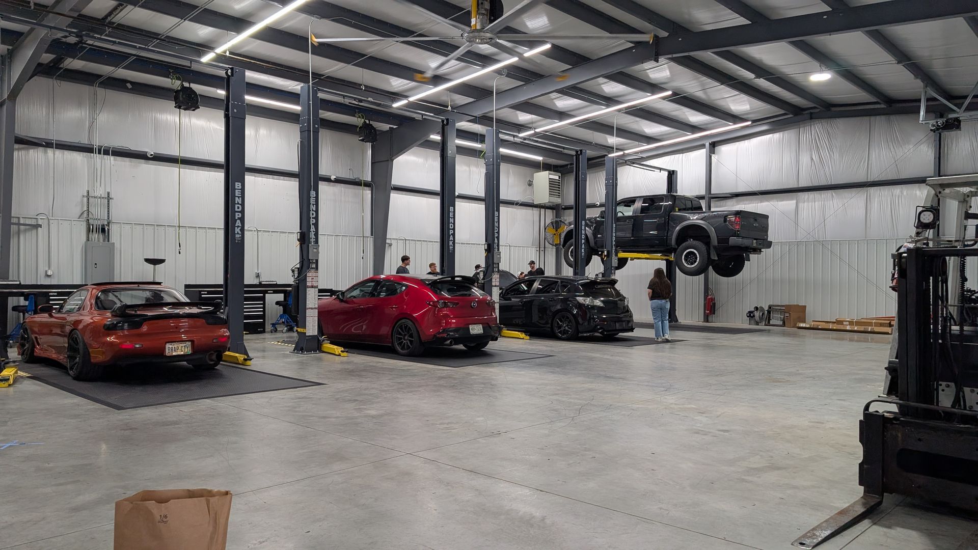Truck lifted on a car hoist inside an auto shop, with hood open and a man partly visible in the foreground.
