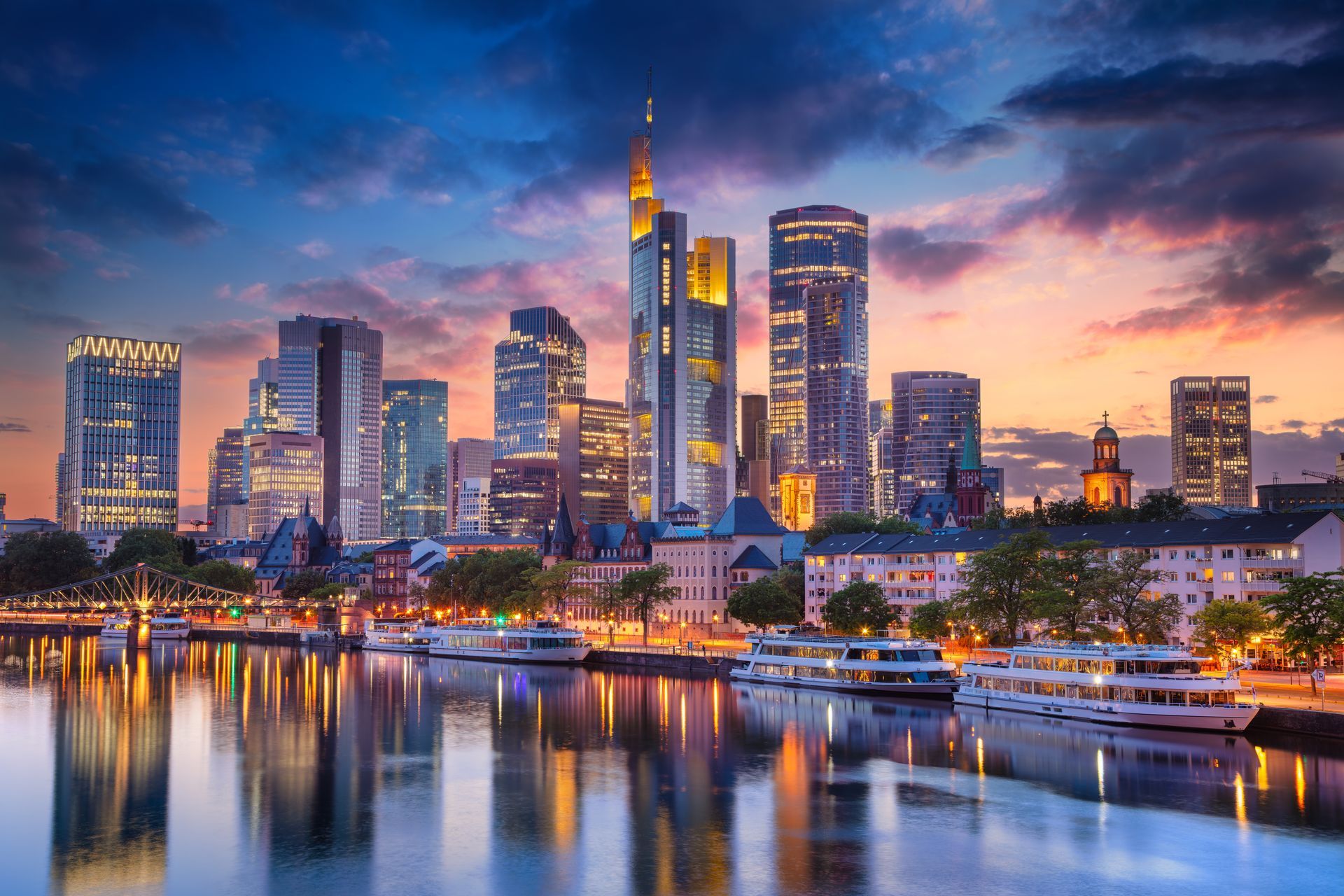Skyline at sunset featuring illuminated skyscrapers reflecting on the Main River with docked boats.