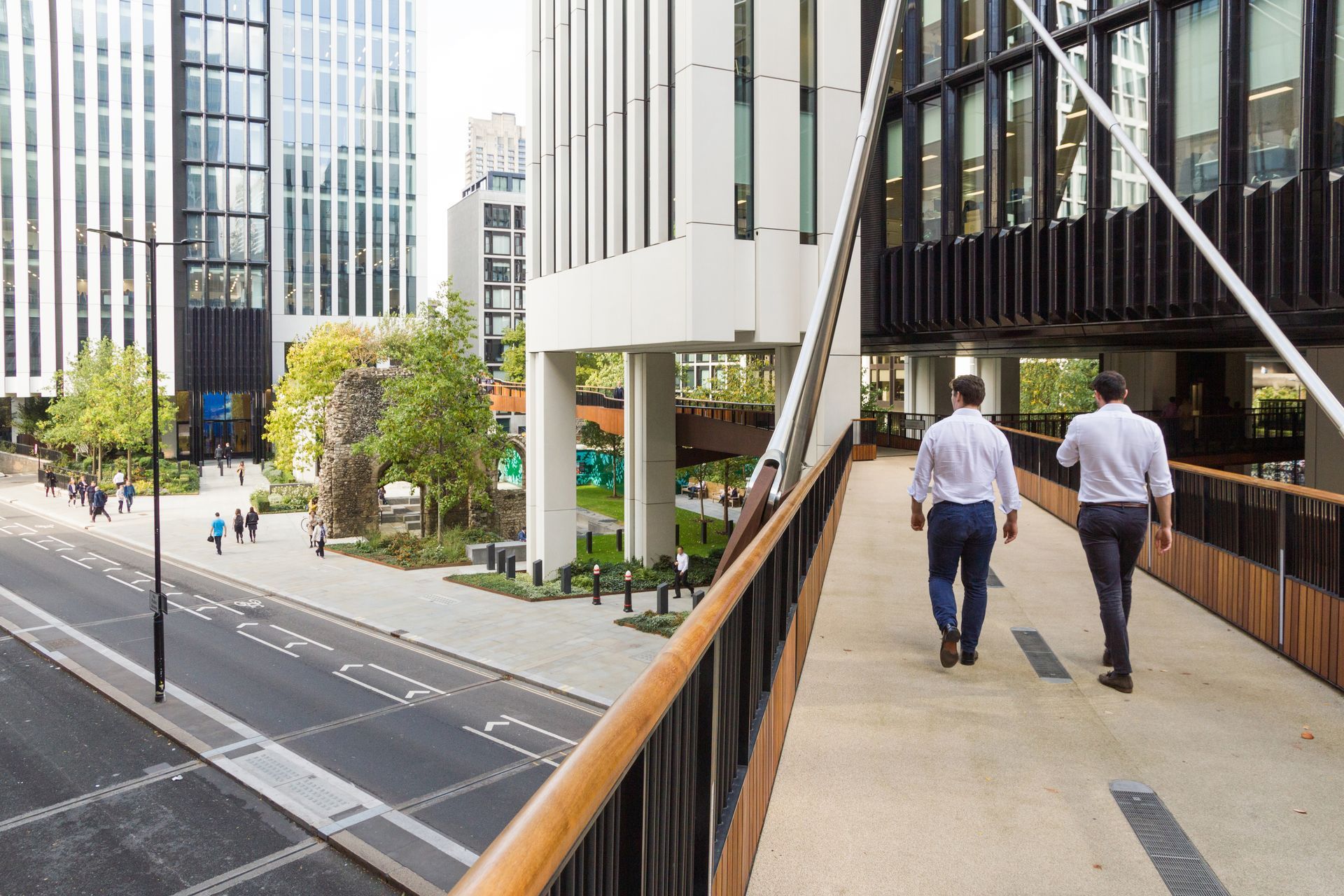 Two people in white shirts and dark trousers walk on an elevated pedestrian bridge next to a modern city street and park.