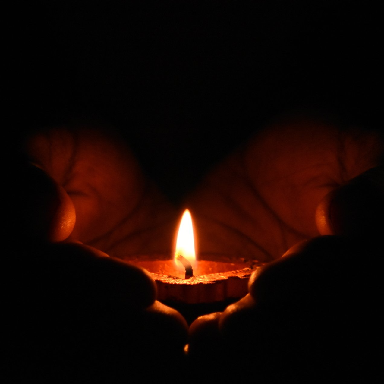 A person is holding a lit candle in their hands in the dark.
