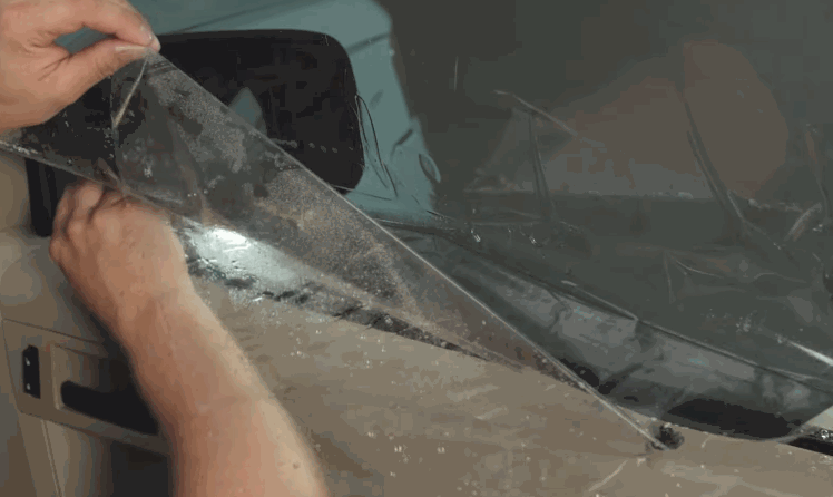 Installer lifts a sheet of window tint against a wet vehicle window while aligning the film along the glass.