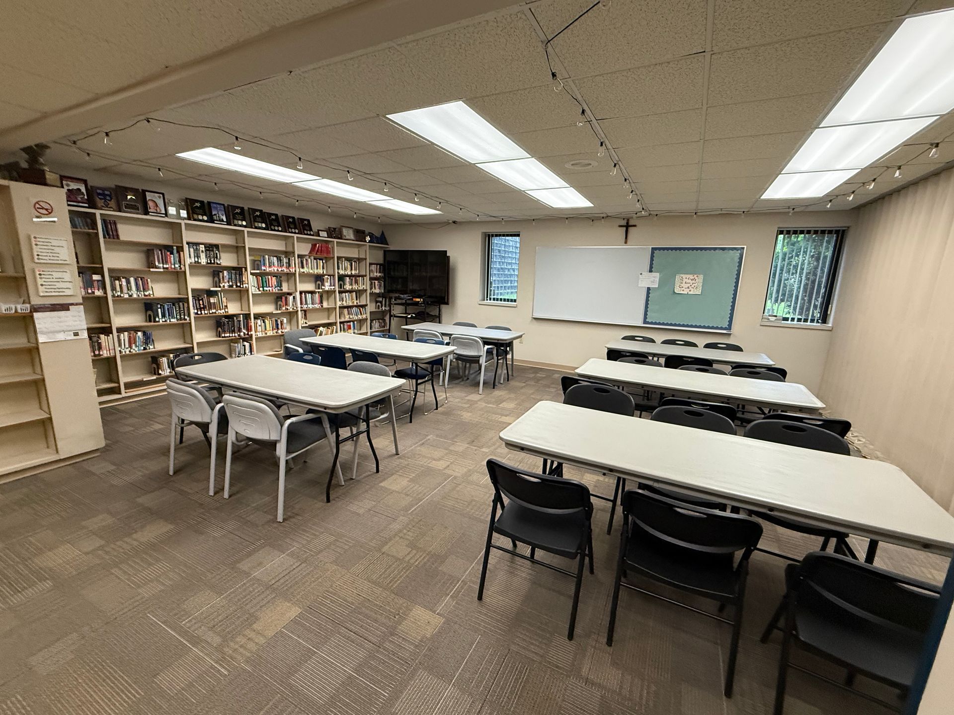 A classroom with tables and chairs, bookshelves, a whiteboard, and windows. The room has beige carpet and fluorescent lighting.