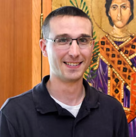 A man wearing glasses is smiling in front of a painting.