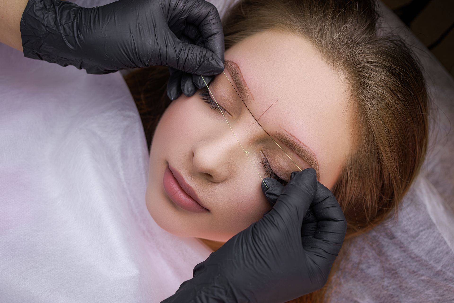 A person with closed eyes has eyebrow mapping with white lines. A gloved hand uses a pencil on their eyebrow.