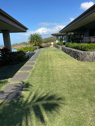 Grassy path between two buildings, with a view of a hillside under a blue sky. A grassy walkway lined with stone pavers connects two building wings under a bright blue sky with tropical landscaping.