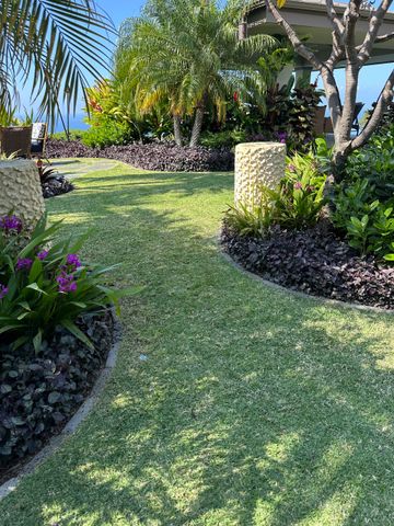 Path through tropical garden with green and purple plants, two stone pillars, and ocean view. A sunny, manicured garden path with purple plants, green lawn, and stone pillars, leading toward a shaded pavilion.