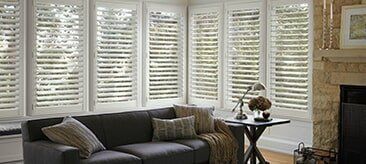 A gray sofa sits in a living room with large windows covered by white plantation shutters and a stone fireplace.