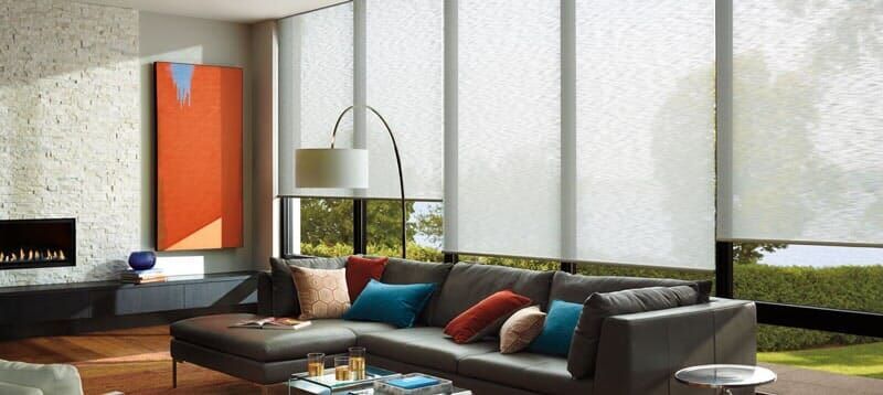 A modern living room with a large gray sectional sofa, an orange art piece, and tall windows covered by roller shades.