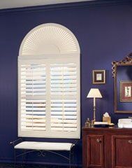 A white arched shutter window installed against a deep purple wall, with a small bench, lamp, and framed art nearby.
