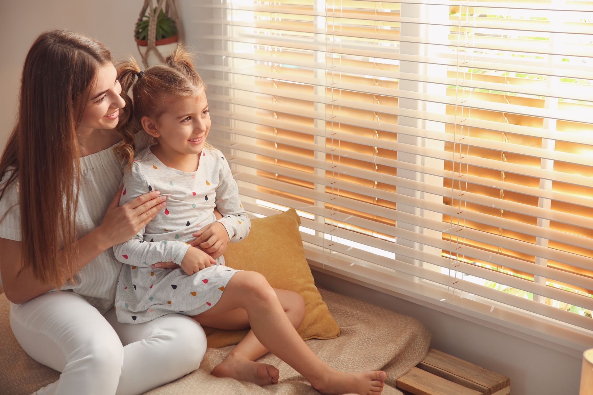 A mother and a young daughter sitting on a window seat beside white horizontal blinds A mother and a young daughter sitting on a window seat beside white horizontal blinds