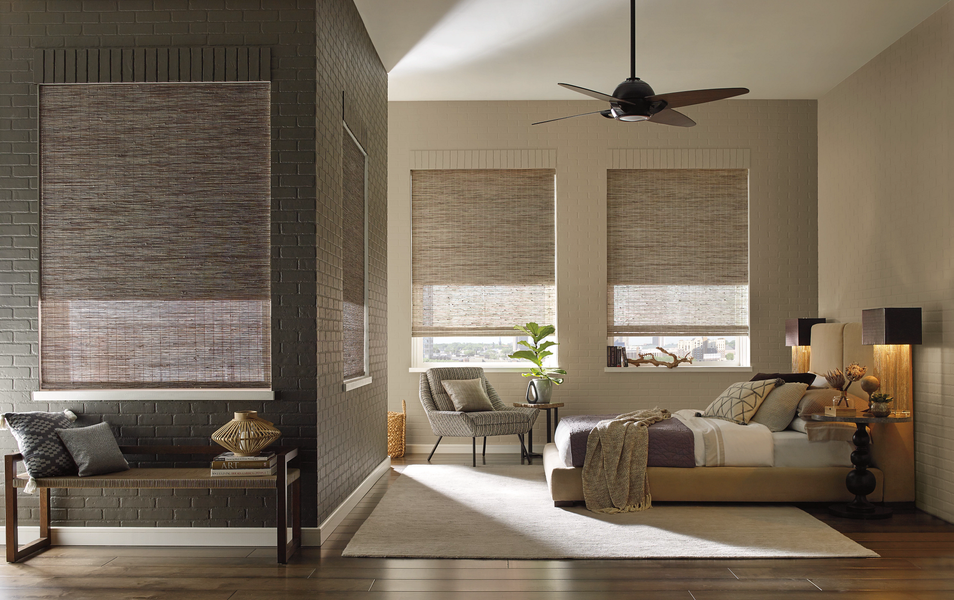 A bedroom with a dark brick wall on the left, two windows with woven shades, a bed, and an armchair.