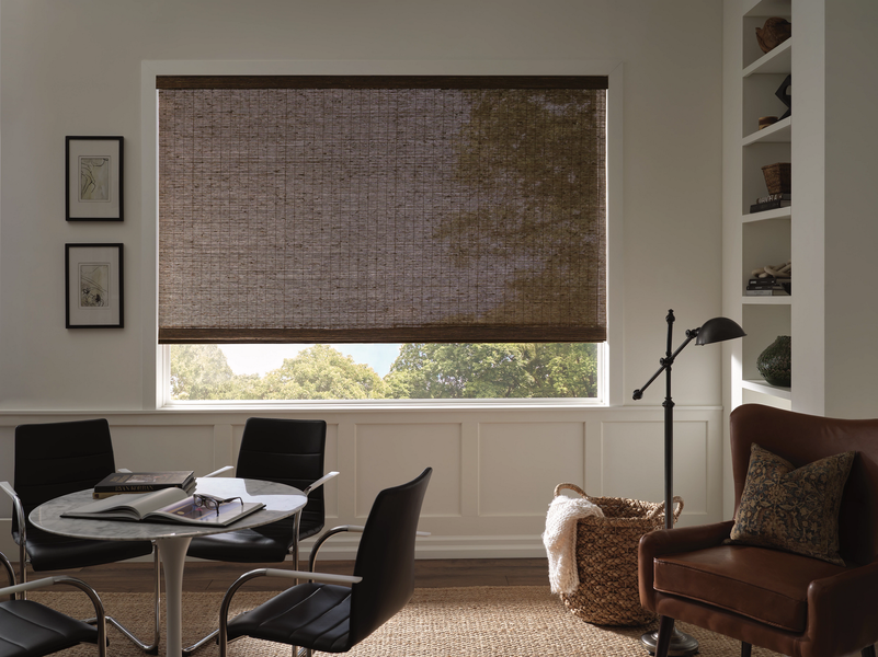 A dining area with a round table, four chairs, a brown leather armchair, and a window covered by a woven bamboo shade.