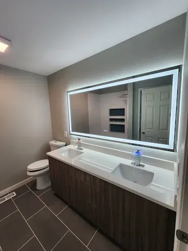 Modern bathroom with double vanity, large backlit mirror, and dark floor tiles.