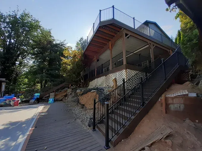Steep walkway and stairs leading to a raised house with a black metal railing and deck on a wooded hillside.