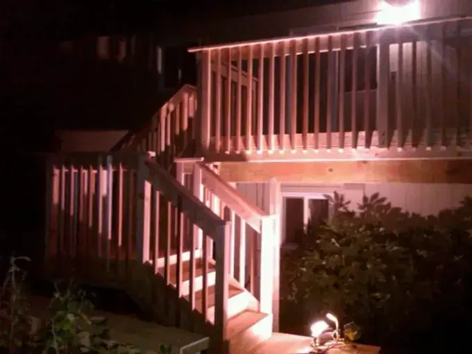 Nighttime exterior of a house with a lit wooden staircase and balcony, with bushes in front