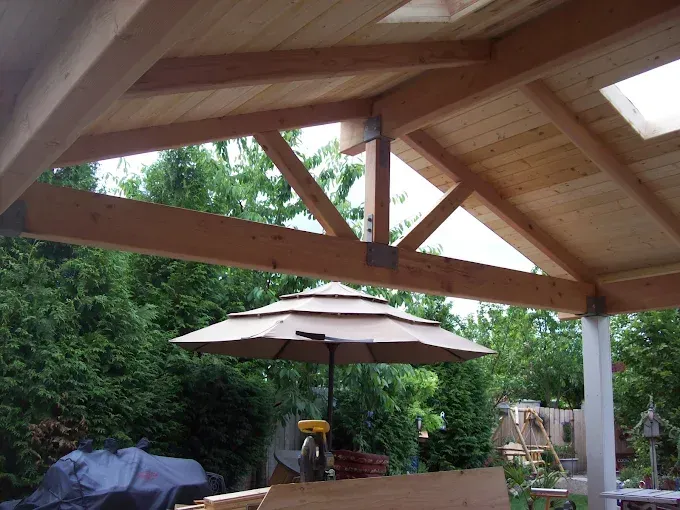 Covered patio with wooden beams, outdoor umbrella, grill, and chairs in a backyard garden
