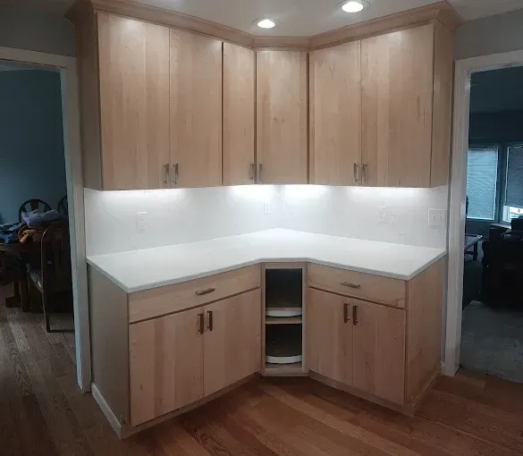 Corner kitchen cabinets with light wood finish, white countertops, and under-cabinet lighting on hardwood floor