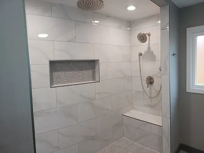 White-tiled walk-in shower with recessed shelf, showerhead, bench, and small window in a gray bathroom.